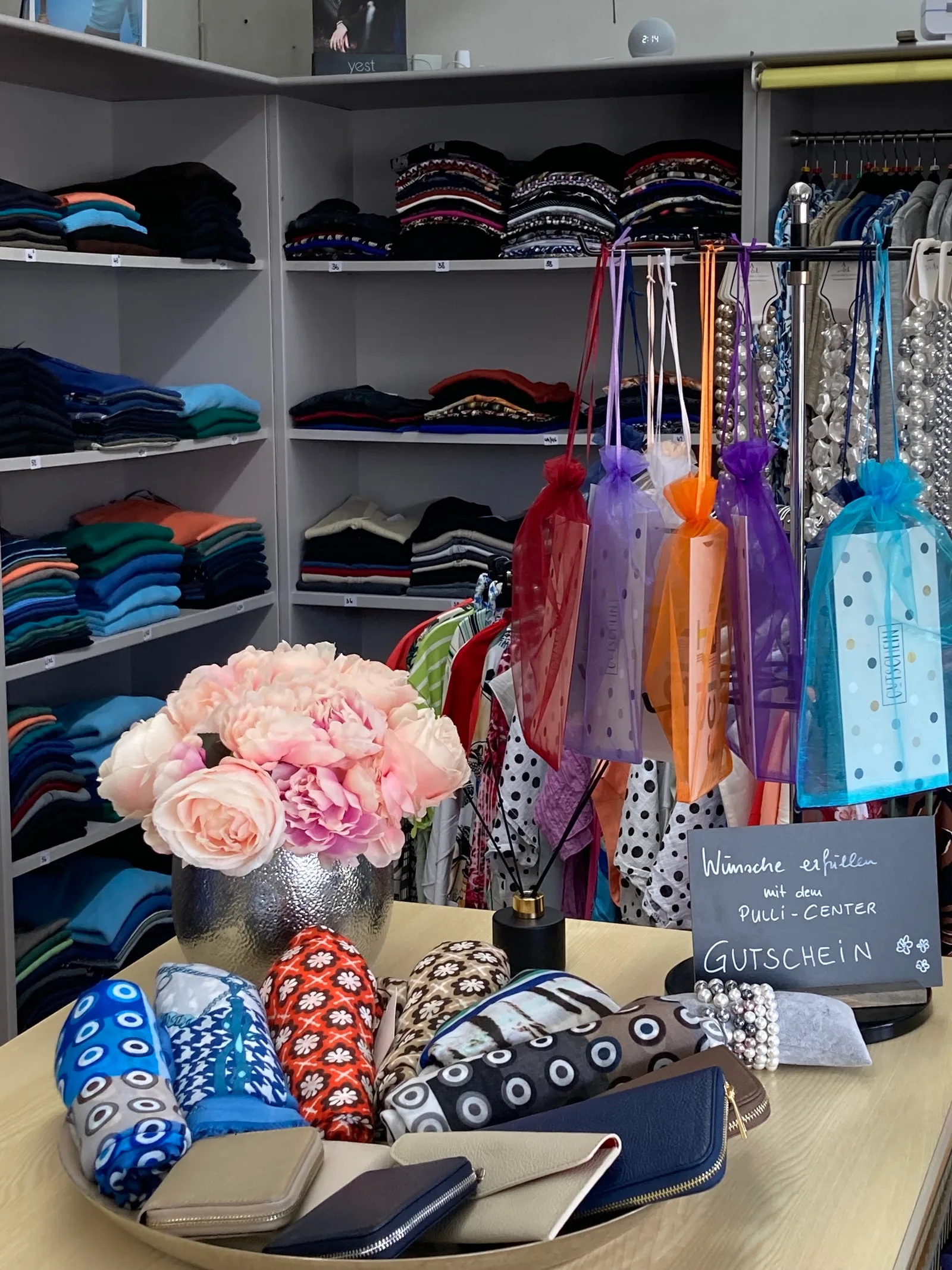 Verkaufstisch mit Seidentüchern, Geldbörsen und Blumen, daneben hängen verpackte Gutscheine in bunten Organzabeuteln vor einer Regalwand
